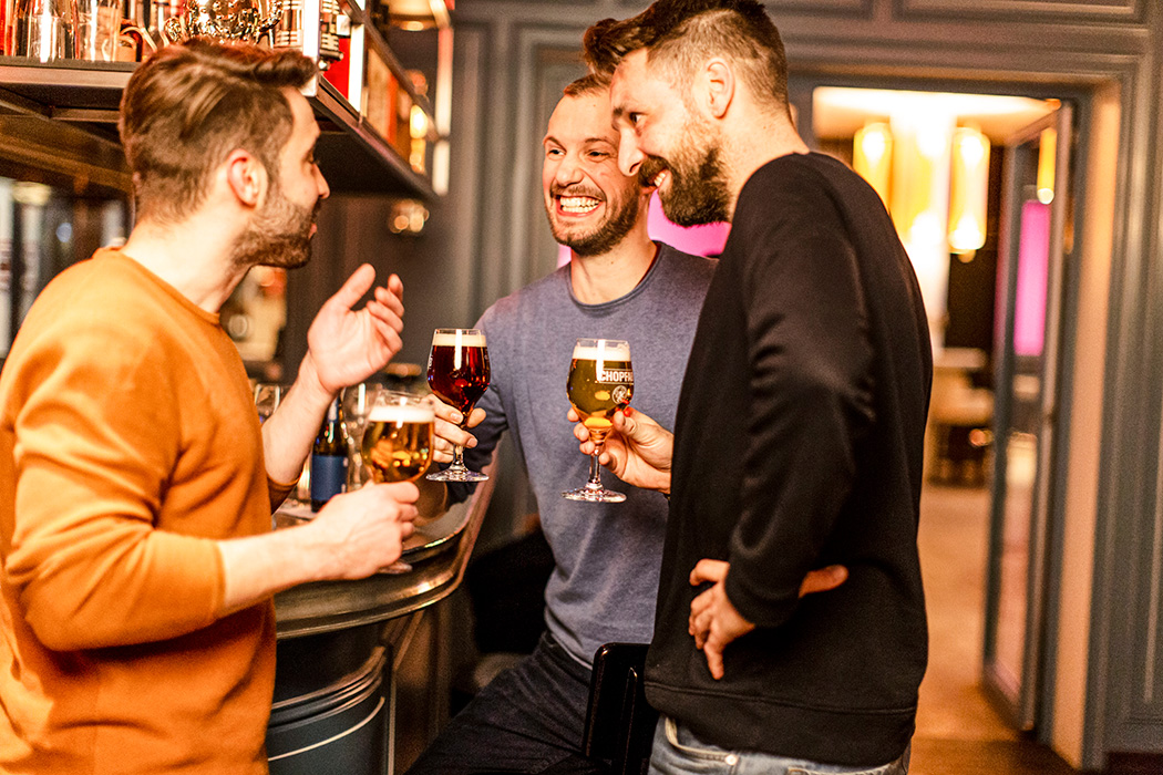 3 Freunde stehen lachend an einer Bar und trinken ein Bier.